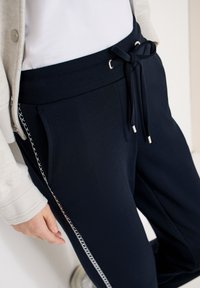Personne portant un pantalon bleu marine avec un détail de couture latérale et une taille à cordon, associé à un haut blanc et un cardigan beige.
