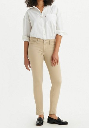 Femme portant un pantalon skinny beige, une chemise blanche boutonnée avec les manches retroussées et des mocassins noirs, debout devant un fond uni.