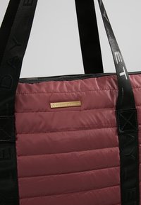 Bourgondische gevoerde draagtas met zwarte riemen, voorzien van een logoplaat in goudkleur. De stof heeft een gladde textuur en een gequilte uitstraling.