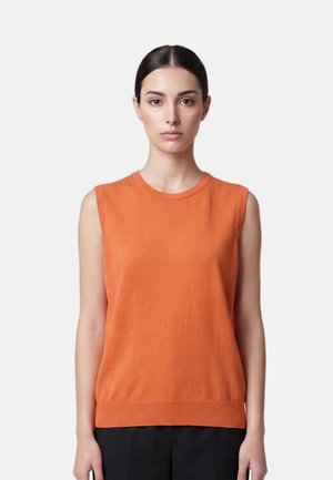 Frau mit dunklen Haaren und Mittelscheitel, die ein oranges ärmelloses Oberteil und schwarze Hose vor einem einfarbigen hellen Hintergrund trägt.