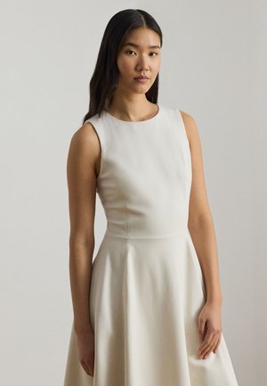 Jeune femme aux longs cheveux foncés portant une robe blanche sans manches, debout devant un fond clair uni.