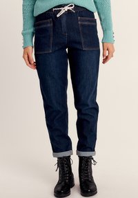 Pantalons en denim foncé avec deux grandes poches avant et une taille à cordon de serrage. Poignets roulés. Associés à des bottes noires à lacets et un pull turquoise.