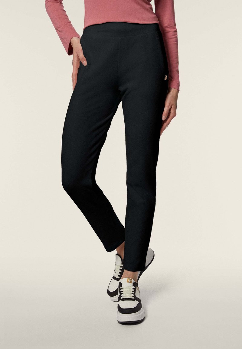 Pantaloni stretch neri con vita alta e vestibilità slim, abbinati a sneakers nere e bianche. Il materiale sembra liscio e flessibile.