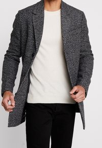 Abrigo de lana texturizado en blanco y negro con cuello con muesca, dos botones y bolsillos laterales, llevado sobre un suéter de canalé crema y pantalones negros.