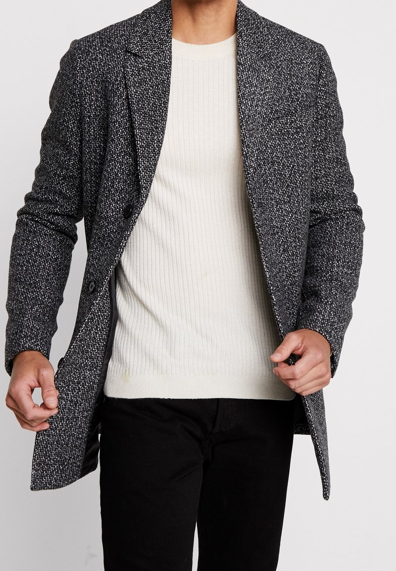 Abrigo de lana texturizado en blanco y negro con cuello con muesca, dos botones y bolsillos laterales, llevado sobre un suéter de canalé crema y pantalones negros.