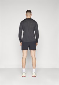 Camisola de manga longa cinza combinada com calções pretos; vista por trás, com um design minimalista e ajuste atlético. Ténis brancos visíveis.