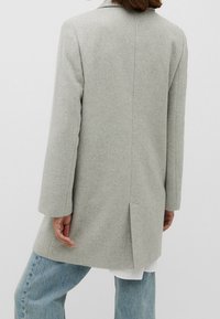 Persona que lleva un abrigo de lana gris claro con una abertura trasera, combinado con jeans azul claro y una camiseta blanca debajo.