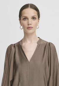 Blusa marrone, testurizzata, con scollatura a V e maniche arricciate. Presenta cuciture a vista lungo il collo, tessuto morbido e silhouette elegante.