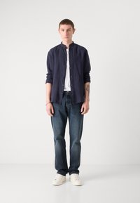 Mørkeblå skjorte med ståkrage, hvit T-skjorte under, mørkeblå jeans og hvite joggesko. Minimalistisk design, enkle detaljer.