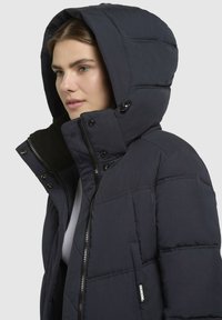 Marineblå pufferjakke med lynlås og trykknaplukning, en stor hætte og quiltet design. Har blødt fleecefor ved kraven og et logo-mærke.