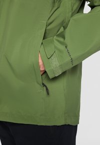 Chaqueta impermeable verde que cuenta con un bolsillo con cremallera, puños ajustables con Velcro y una textura suave. Marca OMNI-TECH en la manga.