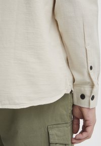 Beige Langarmhemd aus strukturiertem Stoff mit schwarzen Knöpfen an den Bündchen und abgerundetem Saum, kombiniert mit olivgrünen Cargo-Hosen.