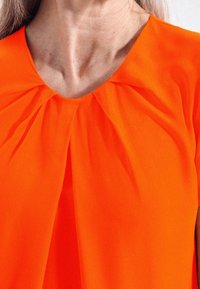 Top arancione brillante con scollatura plissettata. Il tessuto appare leggero e liscio, esaltando il colore vivace e la morbidezza della texture.