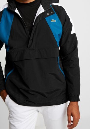 Zwart windjack met een blauw-wit kleurblok ontwerp, lichtgewicht stof, halve ritssluiting aan de voorkant, elastische manchetten en een klein logo accent.