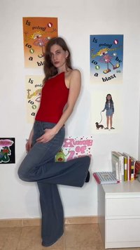 Ung kvinde i rød ærmeløs top og blå jeans poserer med det ene ben bøjet foran en væg dekoreret med farverige tegneserieplakater.