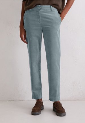 Person wearing light blue straight-fit pants with side pockets and brown slip-on shoes, standing against a plain light background.