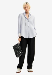 Camicia azzurra chiara con maniche lunghe, pantaloni neri a gamba larga e una borsa nera con pois bianchi, accessoriata con collane sovrapposte.