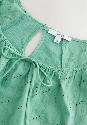 Groene katoen top met een sleutelgat achteraan, gegatherde halslijn en geborduurde patronen met kleine uitsparingen. Merkenlabel zichtbaar aan de binnenkant.