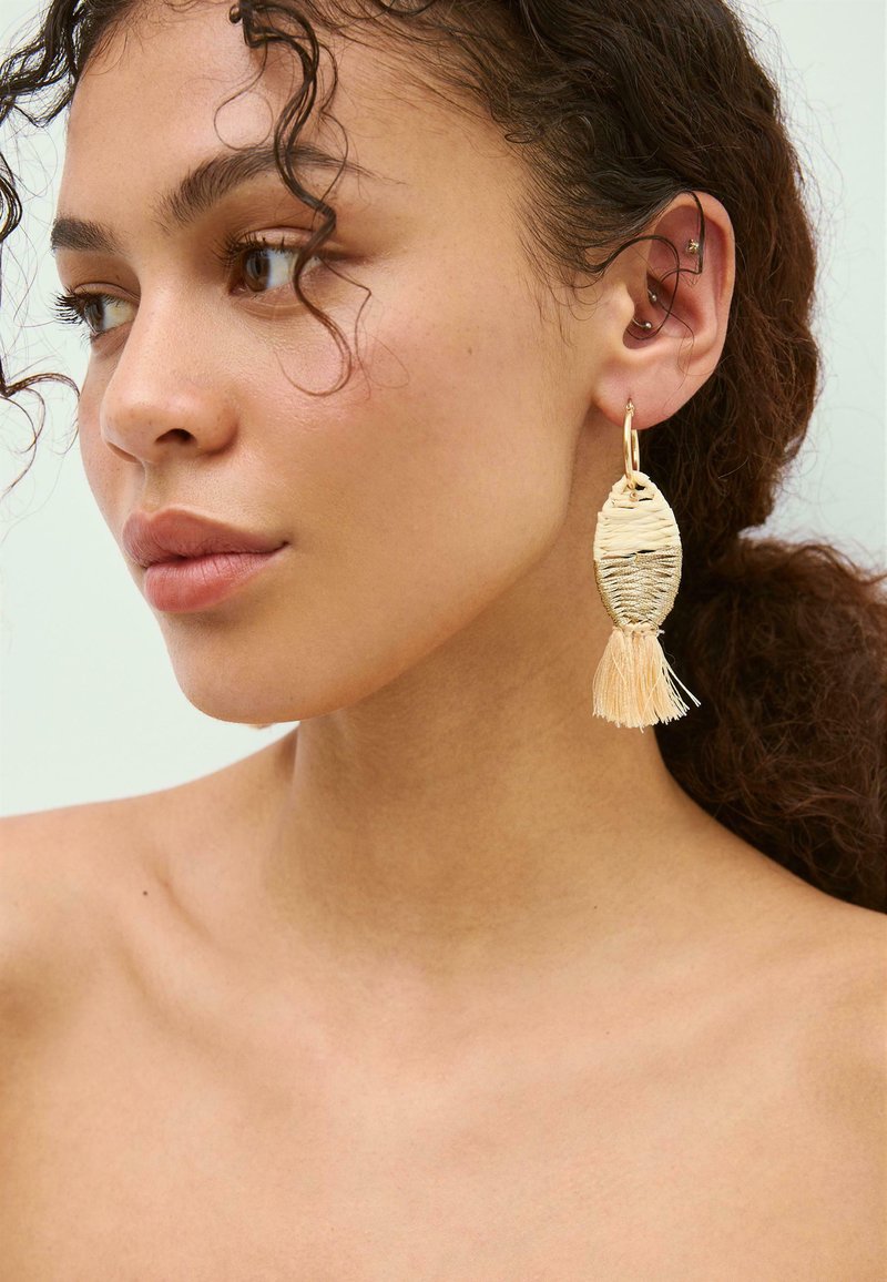 Jeune femme aux cheveux bouclés porte une boucle d'oreille beige tissée à pampilles à l'oreille gauche, montrant plusieurs petits piercings à l'oreille.