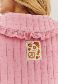 Růžová strukturovaná tkanina s vyvýšenými puntíky a volánkovým límcem; obsahuje smetanovou etiketu s ilustracemi pečiva v teplých barvách.