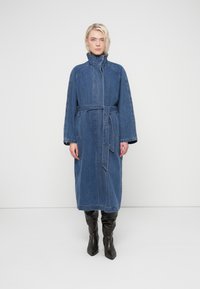 Femme portant un long manteau en jean bleu avec ceinture nouée à la taille, col montant, debout en bottes noires montantes à hauteur de genoux sur fond blanc.