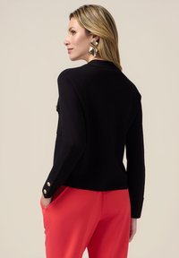 Giacca nera lavorata a maglia con due tasche, dettagli con bottoni dorati sulle maniche, abbinata a pantaloni rosa brillante. Tessuto liscio, design aderente.