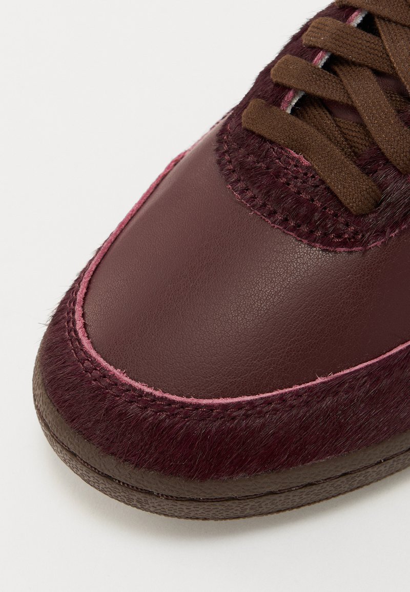 Baskets en cuir bordeaux avec des accents marron texturés et des coutures roses. Bout arrondi et lacets plats. La tige présente des détails en fausse fourrure.
