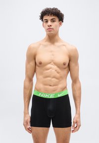 Νέος άνδρας με σγουρά μαλλιά που φοράει μαύρα μποξέρ με πράσινη ζώνη Nike, στέκεται σε απλό φωτεινό φόντο.