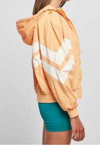 Persikofärgad zip-up jacka med vita chevronränder, åtsittande ärmslut och huva. Slät textur och lättviktig tyg, kombinerad med en teal-botten.