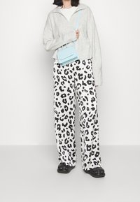 Vrouw met een lichtgrijze trui, zwart-wit luipaardprint wijduitlopende broek, zwarte schoenen en een kleine lichtblauwe schoudertas.