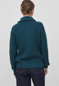 Pull en tricot texturé turquoise avec un large col, des épaules tombantes et un ourlet côtelé, associé à un jean en denim foncé. Vue de dos présentée.