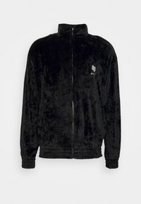 Zwarte jas met ritssluiting gemaakt van zachte, pluizige stof. Heeft een hoge kraag, elastische manchetten en een klein geborduurd logo op de borst.