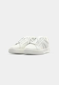 White leather sneakers with a smooth texture, lace-up front, and rubber sole featuring a diamond pattern. Side logo accent is present.