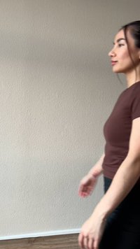 Žena v hnědém tričku a černých kalhotách jde uvnitř kolem béžové texturované zdi a dřevěné podlahy.