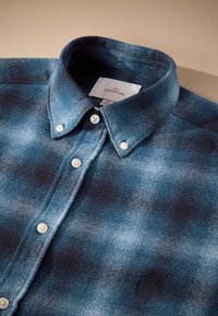 Chemise à carreaux bleue et grise avec boutons blancs et col boutonné, posée à plat sur une surface claire.