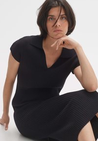 Abito nero a costine con scollo a V e maniche corte, caratterizzato da un colletto strutturato e una silhouette aderente. Finitura liscia.