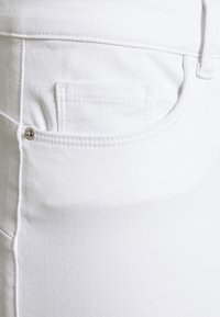 Tela de mezclilla blanca que presenta un bolsillo delantero con costuras visibles y un detalle de botón metálico plateado. Textura suave y diseño ajustado.