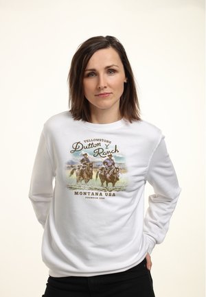Vrouw met bruin haar die een wit sweatshirt draagt met twee cowboys te paard en de tekst "Yellowstone Dutton Ranch, Montana USA."