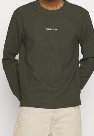 Sudadera de manga larga en verde oliva con tejido texturizado, cuello redondo y logo "CALVIN KLEIN" en blanco en el pecho. Diseño sencillo con acentos de canalé.
