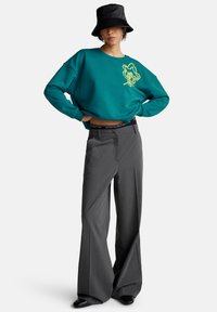 Felpa teal con grafica gialla, vestibilità comoda, abbinata a pantaloni grigi a vita alta e cappello a secchiello nero, con cintura con marchio.