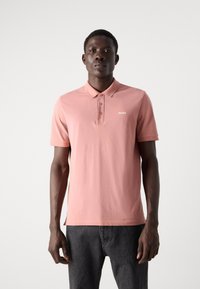 Polo à manches courtes en coton rose clair, avec un col, une patte de boutonnage à trois boutons et un petit logo blanc sur la poitrine.