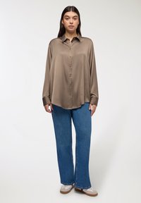 Camicia satinata oliva, oversize e a maniche lunghe, con bottoni davanti; abbinata a jeans a gamba larga azzurri e sneakers marroni.
