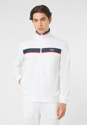 Młody mężczyzna stoi ubrany w biały rozpinany na zamek kurtkę z granatowo-czerwonymi poziomymi paskami i logo FILA na piersi, ręce w kieszeniach.