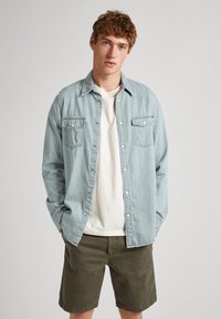 Camicia di denim blu chiaro con chiusura a bottoni e due taschini sul petto, indossata sopra una maglietta bianca, abbinata a pantaloni corti verde oliva.