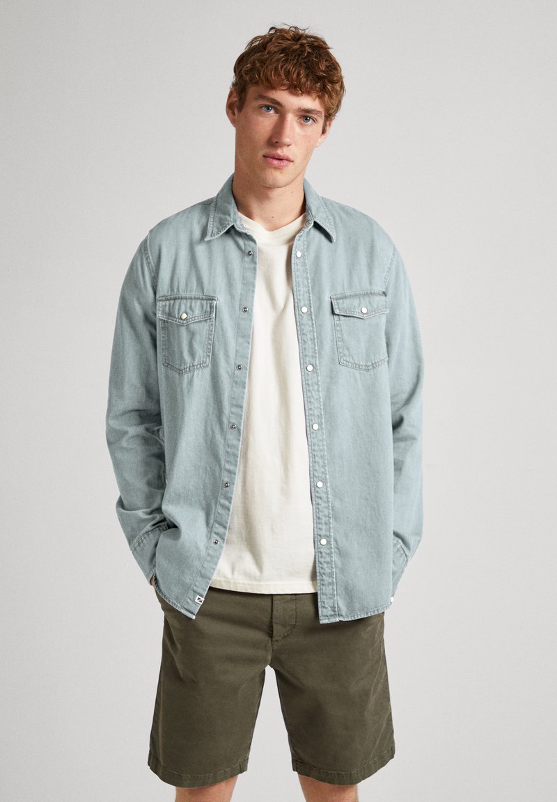 Camicia di denim blu chiaro con chiusura a bottoni e due taschini sul petto, indossata sopra una maglietta bianca, abbinata a pantaloni corti verde oliva.