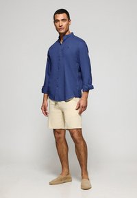 Granatowa koszula z długim rękawem i kołnierzykiem zapinanym na guziki, w zestawieniu z beżowymi szortami i neutralnymi espadrylami. Gładki materiał, klasyczny krój.