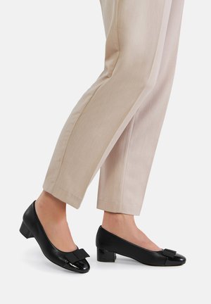Czarne niskie buty na obcasie ze skóry z błyszczącym czubkiem i małą kokardką. Noszone z beżowymi spodniami, ukazujące gładką fakturę.