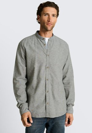 Man wearing a light grey button-up shirt with a mandarin collar and blue jeans, standing against a plain light background.