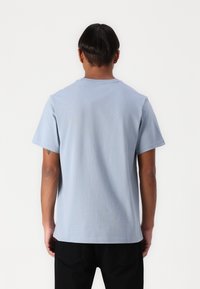 Tricou din bumbac de culoare albastru deschis, cu mâneci scurte, guler rotund și o croială lejeră. Vedere din spate care evidențiază o textură uniformă și detalii subtile de cusătură.