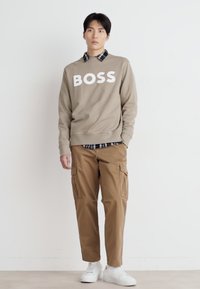Sudadera beige con cuello redondo con "BOSS" en grandes letras blancas, superpuesta a una camisa a cuadros, combinada con pantalones cargo ligeros marrones y zapatillas blancas.
